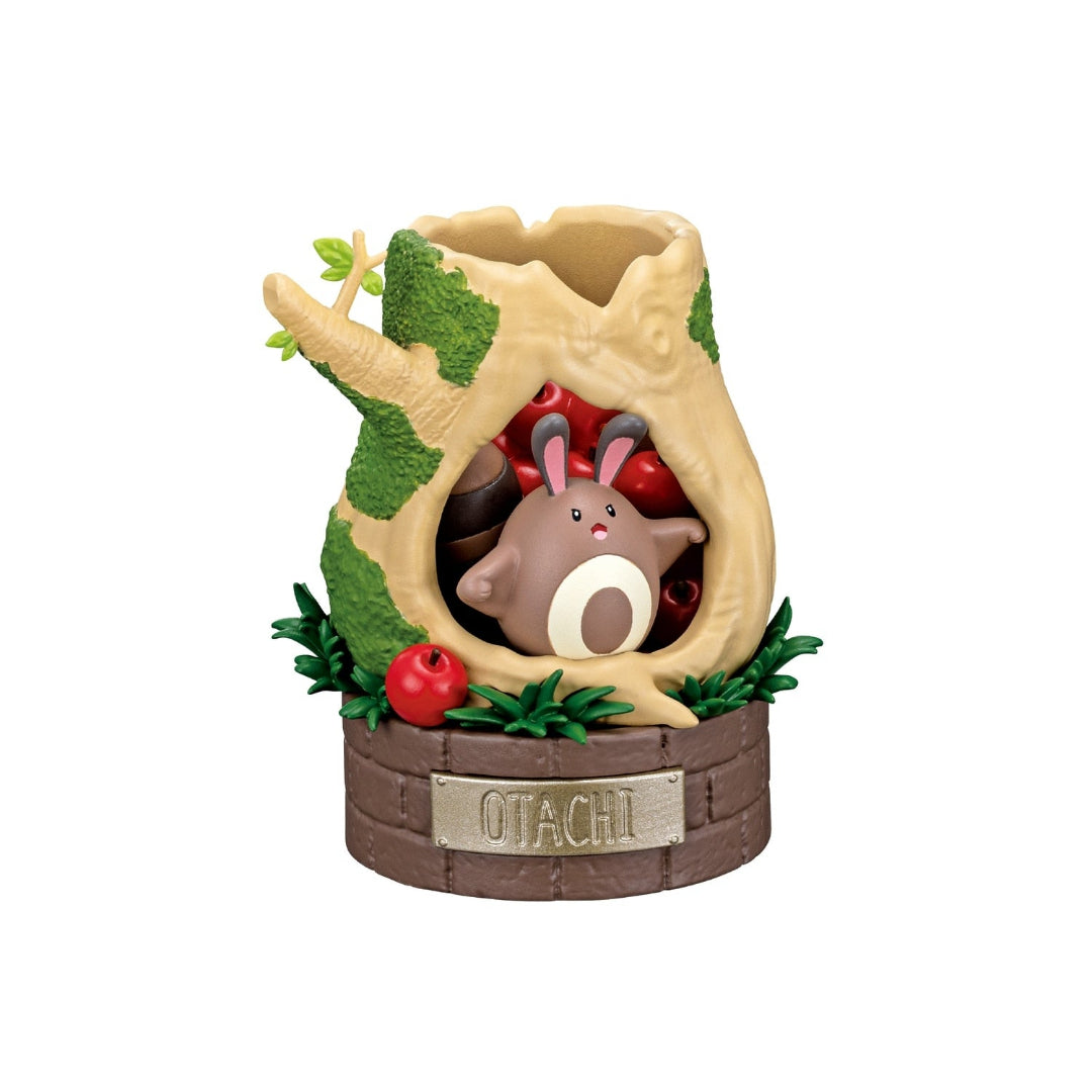 A small Pokémon figure featuring Sentret sitting inside a hollow tree stump filled with red apples, surrounded by green leaves. The display rests on a round brick-patterned base labeled “OTACHI.”
